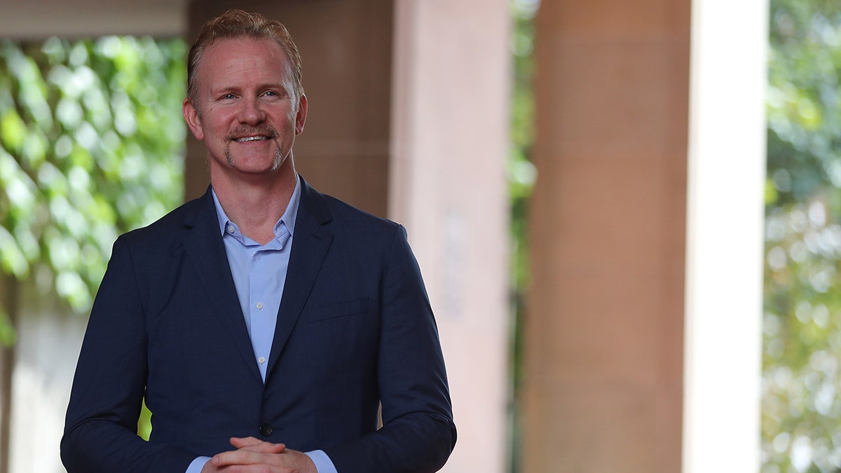 Morgan Spurlock in a navy suit and light blue shirt smiles 