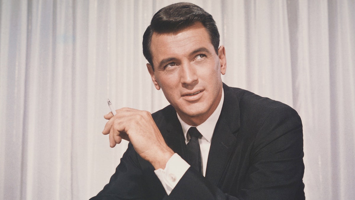 A close-up of Rock Hudson holding a cigarette and wearing a suit