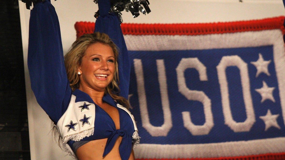 A Dallas Cowboys Cheerleader delivering cheer in front of a USO sign