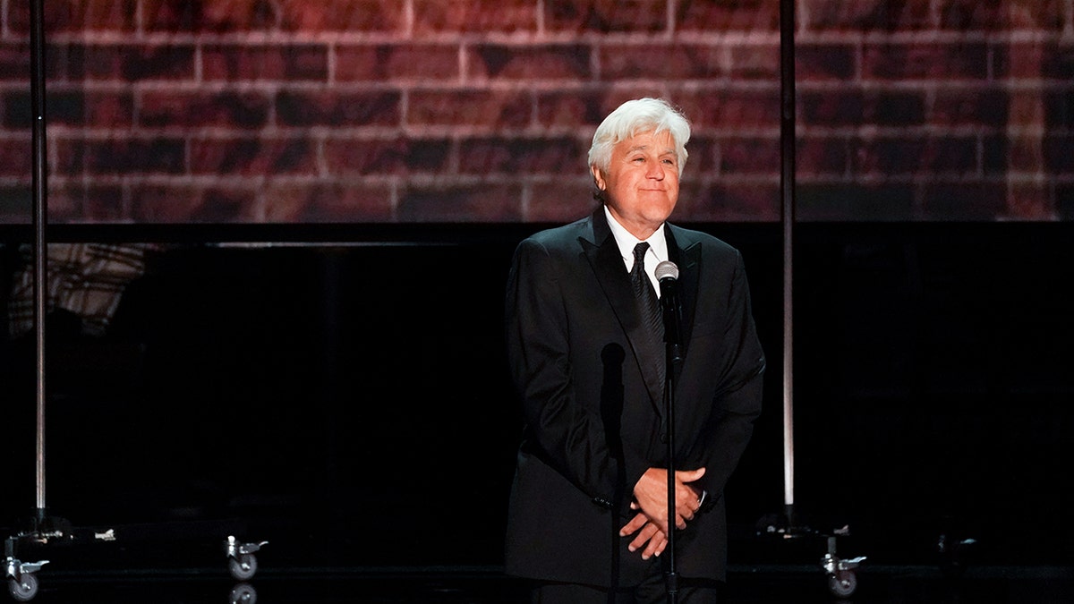 Jay Leno standing on stage in front of a brick wall