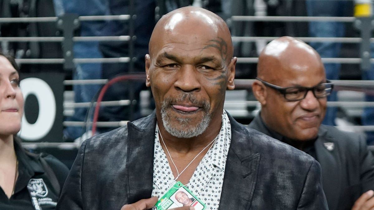 Former heavyweight boxing champion Mike Tyson wears a dark jacket and white shirt as he stands on the field before the NFL football game between the Las Vegas Raiders and the Pittsburgh Steelers in Las Vegas.