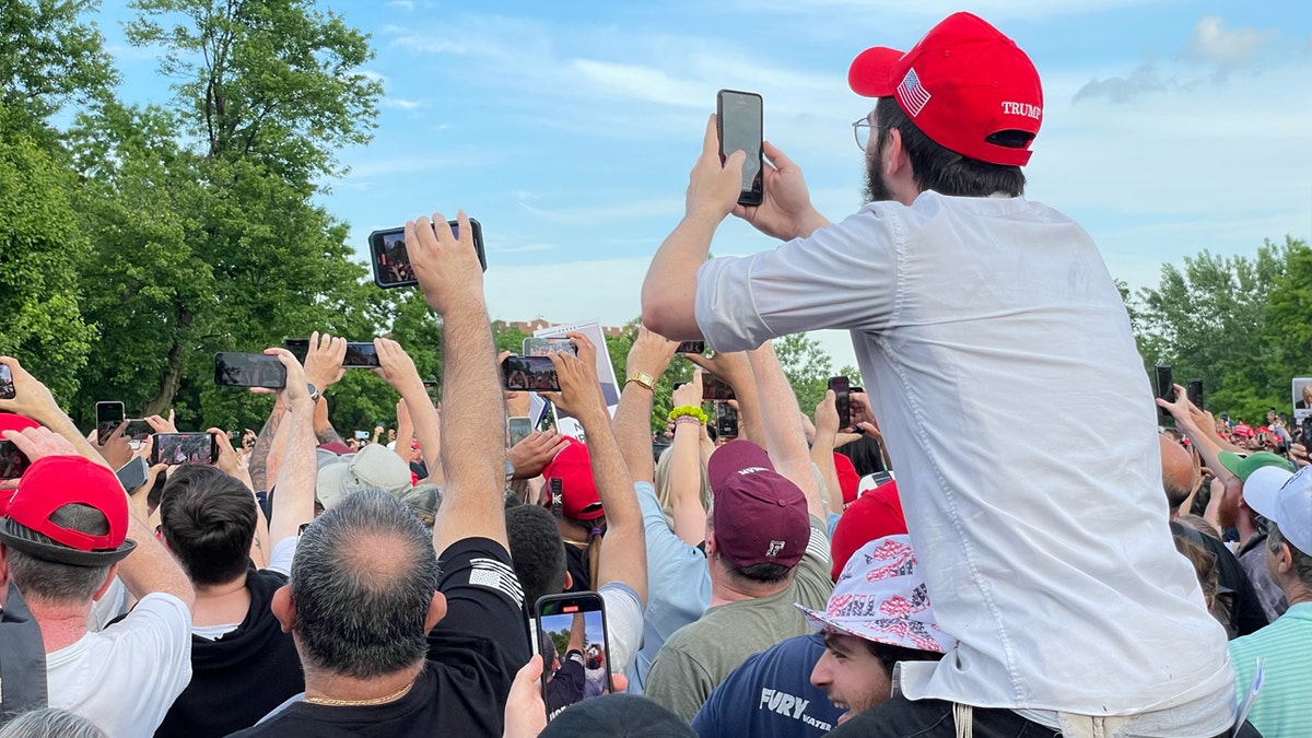 A man in white shirt and red pro-Trump hat sits on another man's shoulders while trying to snap a photo of the former president