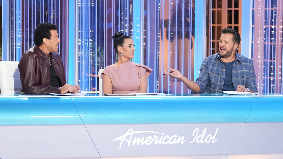 Lionel Richie in a dark purple/brown leather jacket sits behind the judge's desk with Katy Perry in the middle in pink and Luke Bryan at the other end in a plaid shirt