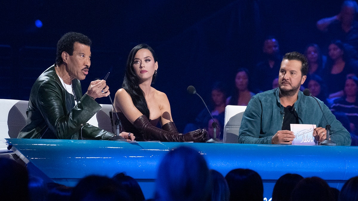 Lionel Richie habla con un bolígrafo sentado detrás de la mesa del juez en "American Idol" con Katy Perry a su lado y Luke Byran mirándole