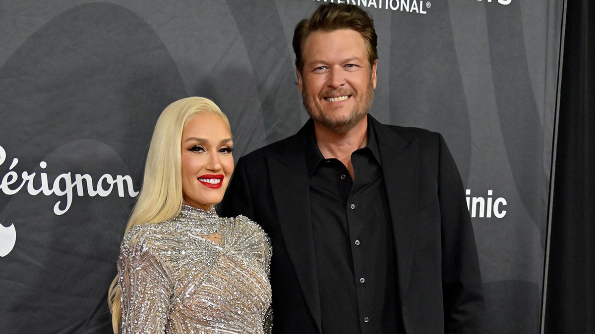 Gwen Stefani in a silver jeweled dress smiles on the carpet with husband Blake Shelton in a black suit and shirt