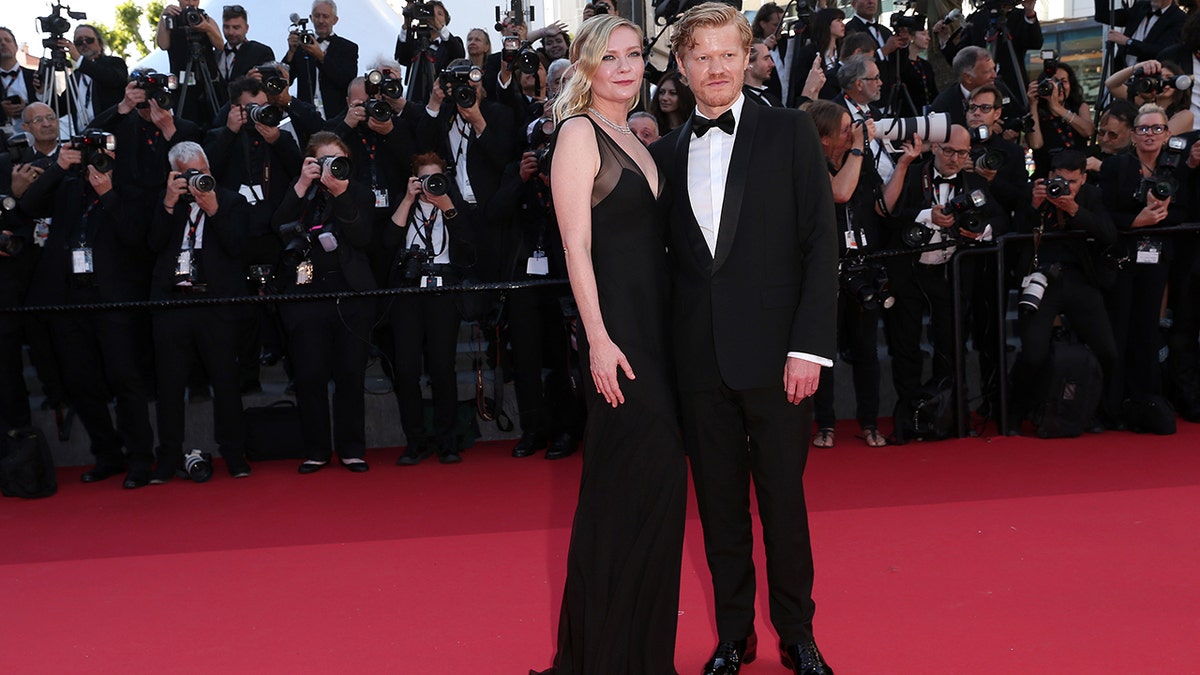 Kirsten Dunst e Jesse Plemons no tapete vermelho do Festival de Cinema de Cannes de 2024 usando um vestido preto com um laço no cabelo e um smoking preto.