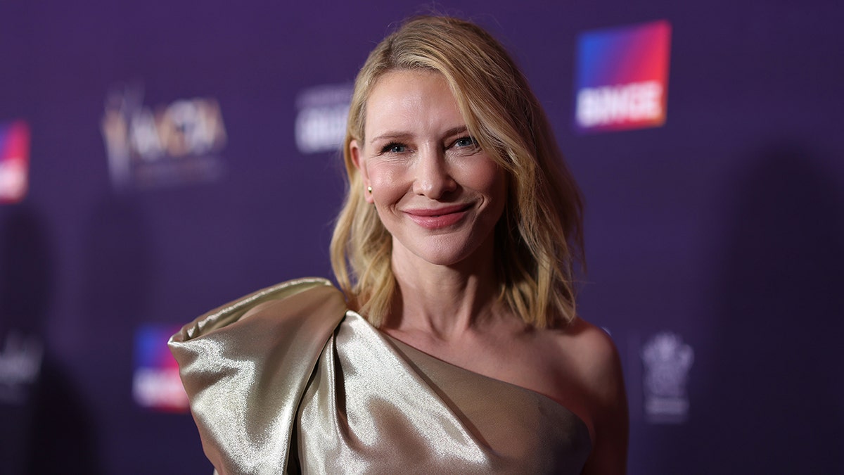 Kate Blanchett in a gold one shoulder dress with a bow smiles on the carpet