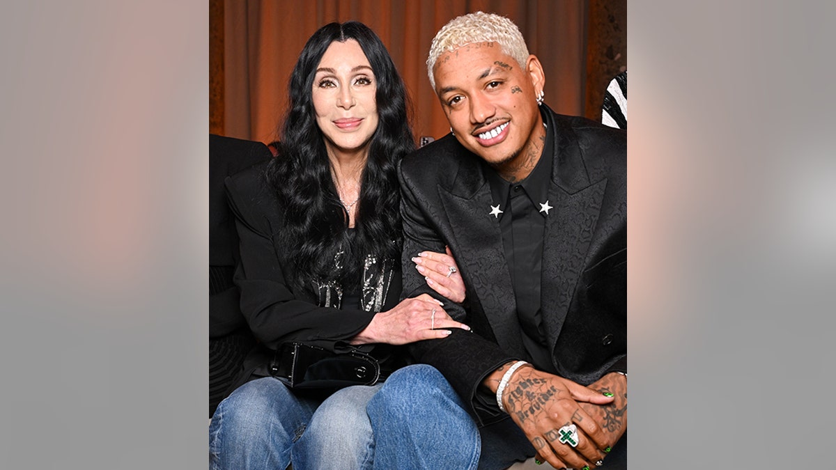 Cher in a black top and jeans holds on to boyfriend Alexander Edwards at the Balmain show