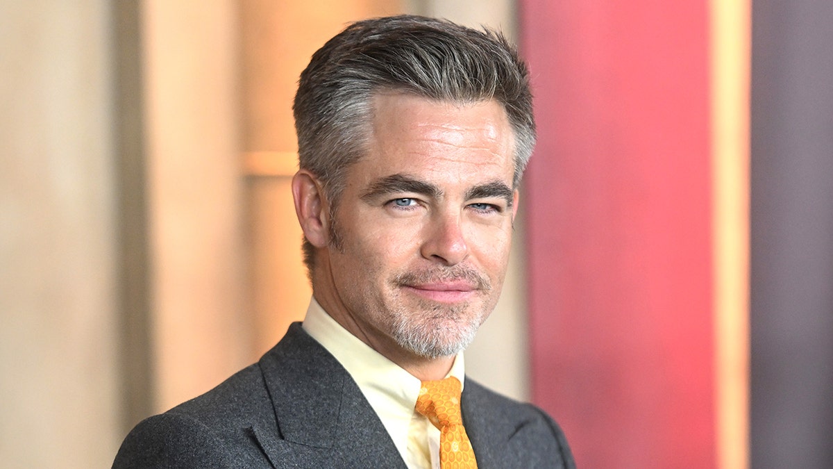 Chris Pine smirks on the carpet in a grey suit, yellow shirt and yellow/orange tie