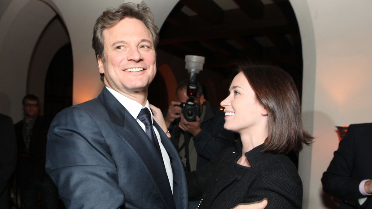 Colin Firth in a dark suit looks slightly over his shoulder as Emily Blunt in black looks up at him lovingly