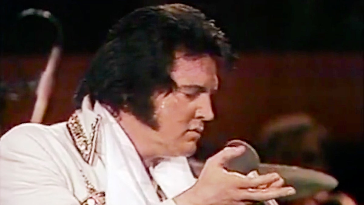 Elvis Presley in a white jacket adorned with a design holds onto his microphone passionately 