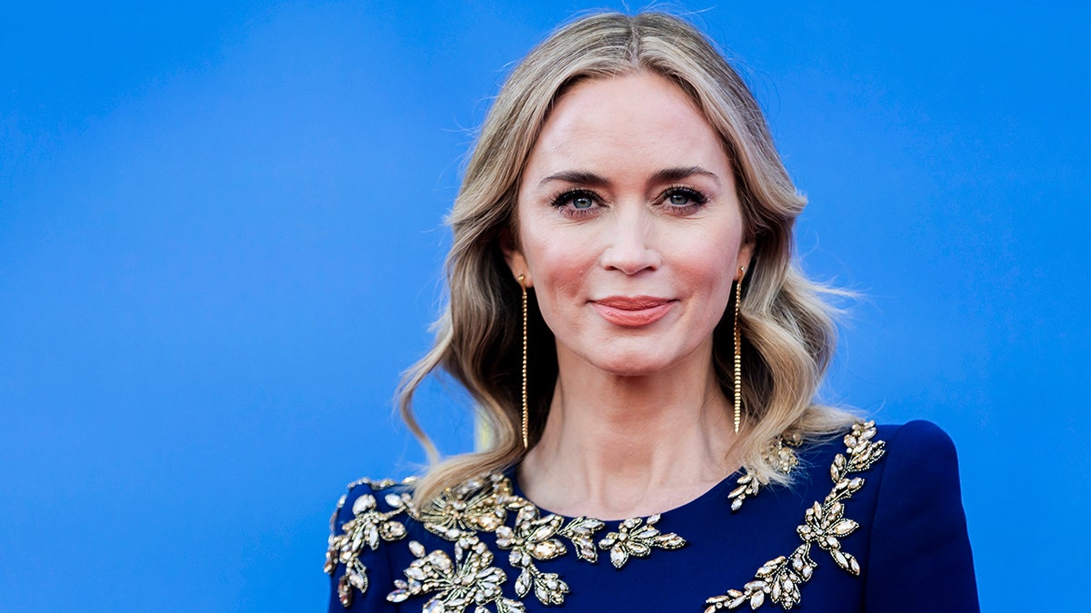 Emily Blunt soft smiles on the carpet in a dark blue dress with a pattern