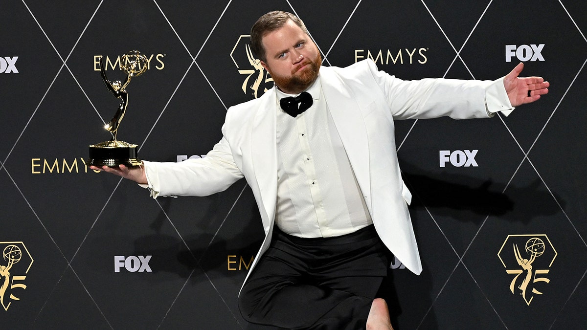 Paul Walter Hauser in a white tuxedo jacket and black pants at the Emmy's makes a silly face and jumps in the air holding his award