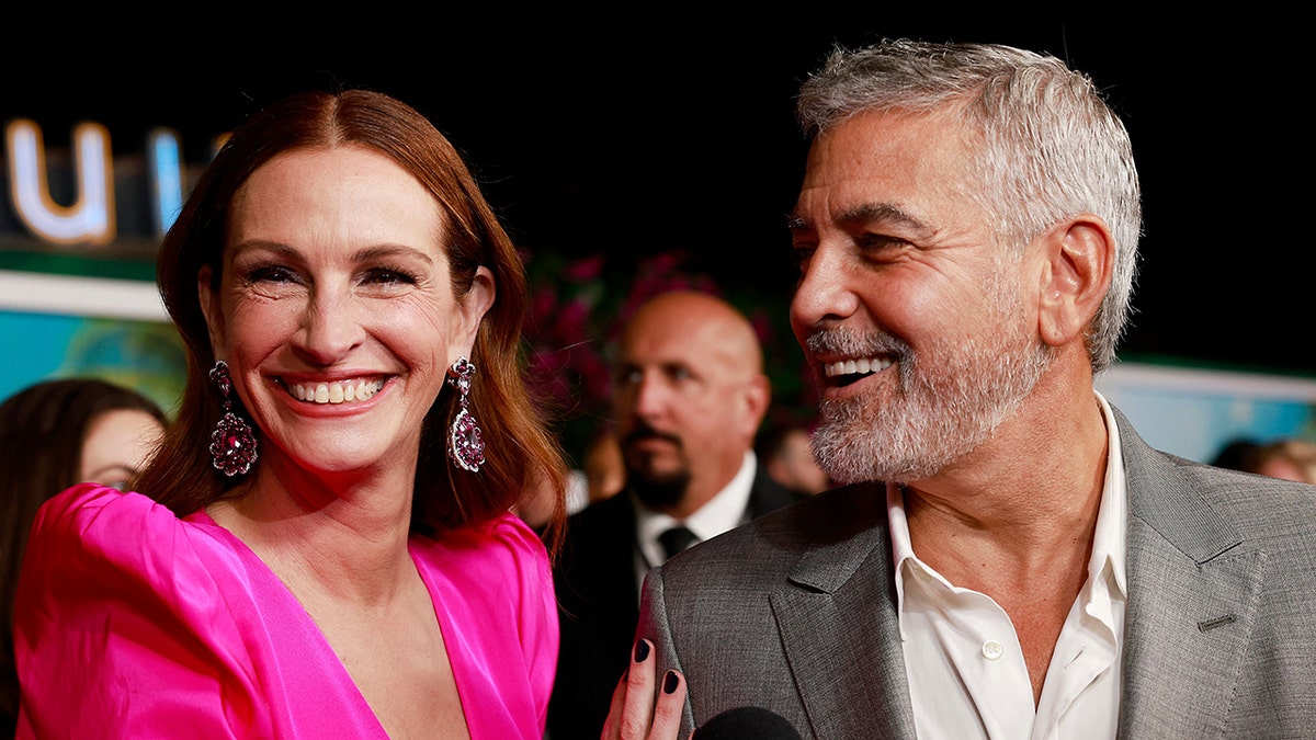 Julia Roberts in a hot pink dress smiles on the carpet as George Clooney in a grey suit smiles and looks at her