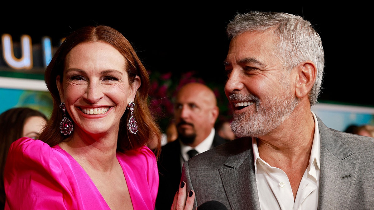 Julia Roberts con un vestido rosa picante sonríe en la alfombra mientras George Clooney con un traje gris sonríe y la mira