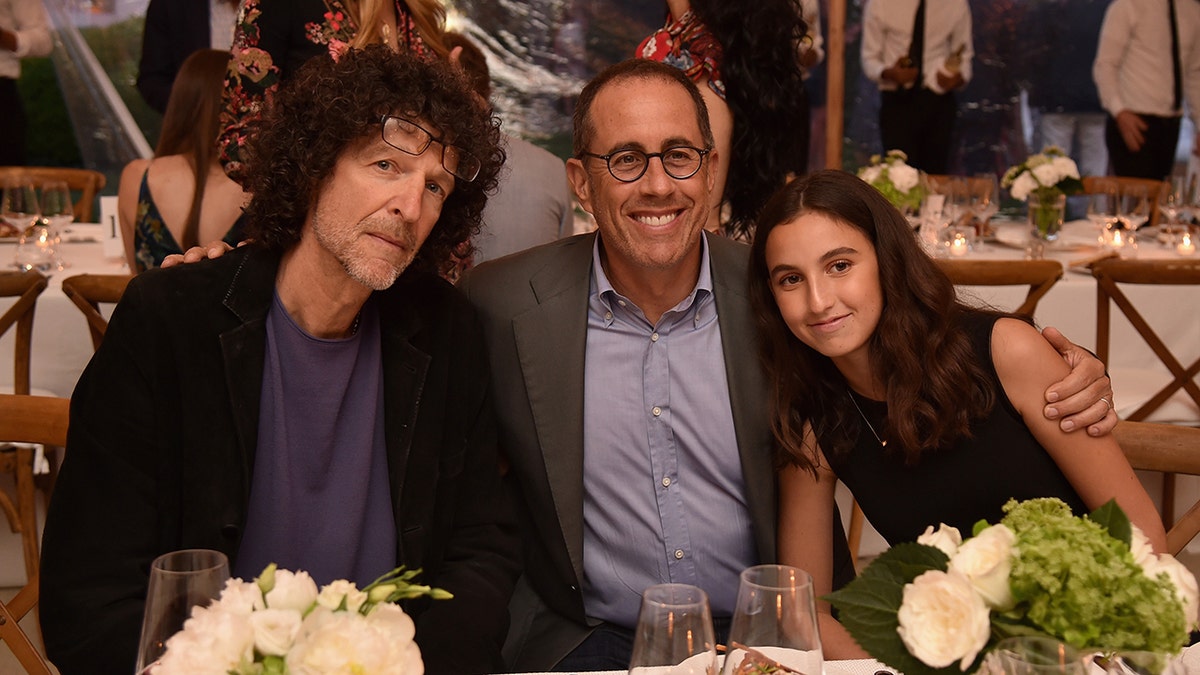 Howard Stern con chaqueta negra se sienta junto a Jerry Seinfeld con chaqueta gris y su hija Sascha en un acto en Nueva York