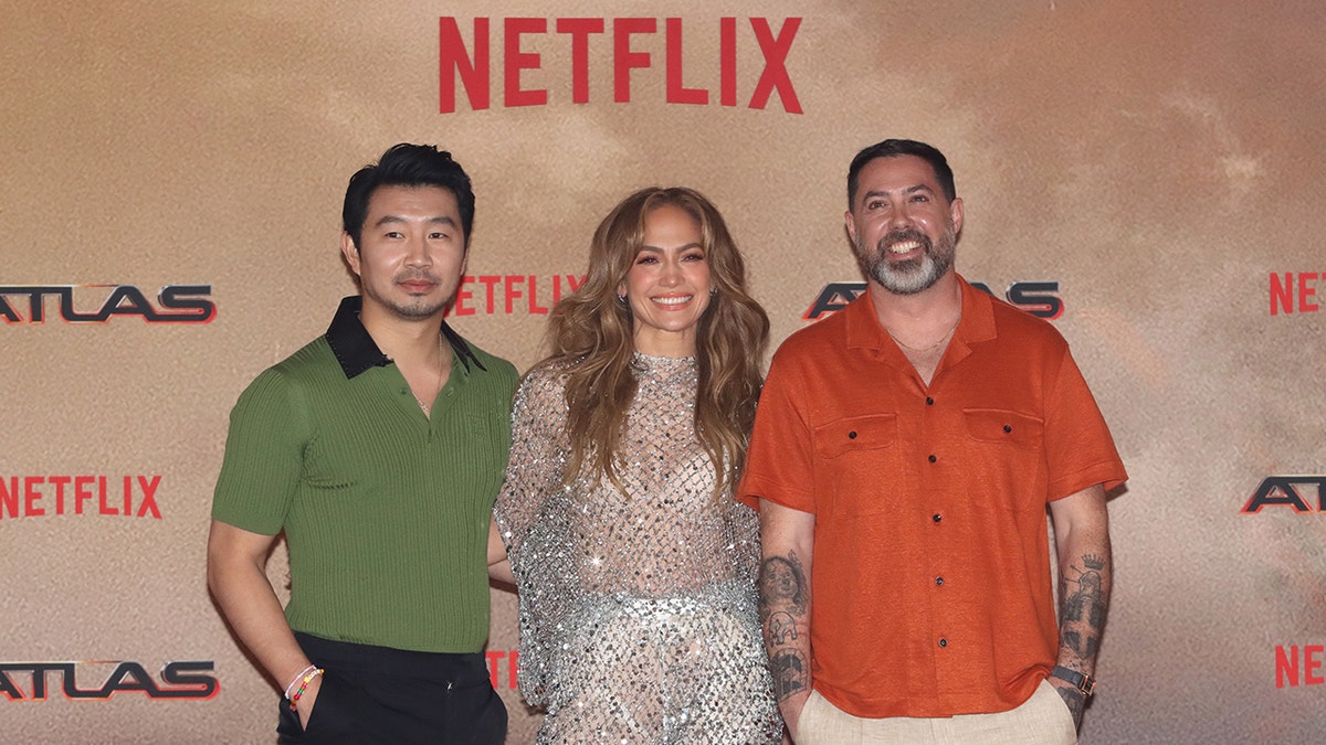 Simu Liu in a green collar shirt stands next to Jennifer Lopez in a sparkly jumpsuit next to Brad Peyton in an orange linen shirt on the carpet
