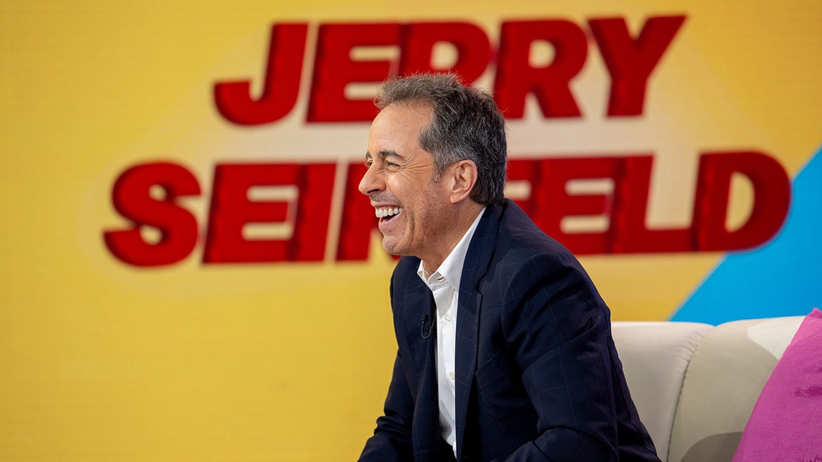 Jerry Seinfeld laughs in a black suit with his name in red letters behind him on TODAY