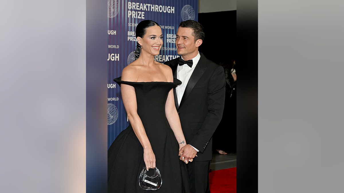 Katy Perry in a black off the shoulder gown holds hands with Orlando Bloom in a tuxedo who lovingly looks at her