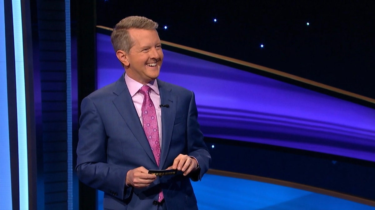Ken Jennings in a blue suit, light pink shirt and dark pink tie laughs on stage of "Jeopardy! Masters"