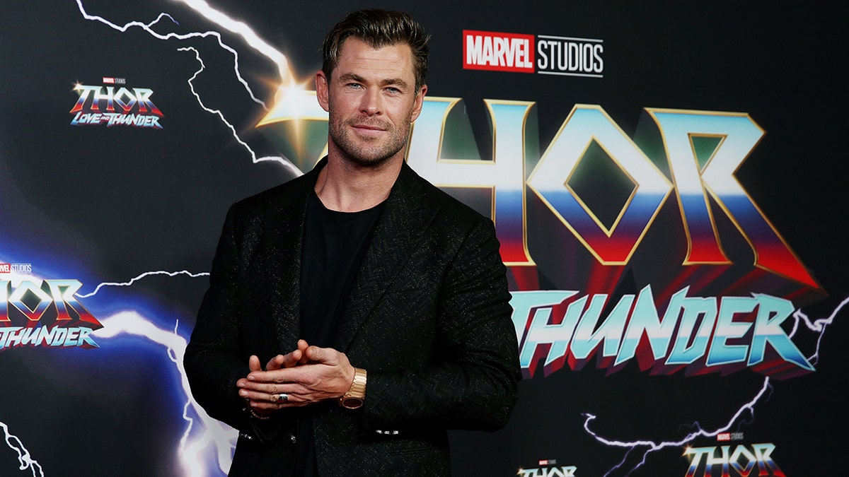 Chris Hemsworth in a black shirt and jacket puts his hands together on the carpet for a Marvel movie premiere