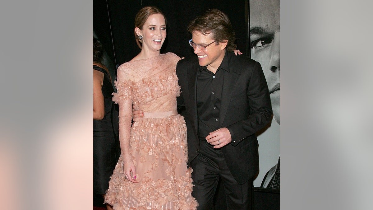 Emily Blunt in a peach ruffled dress laughs with Matt Damon in a black suit and shirt and eyeglasses