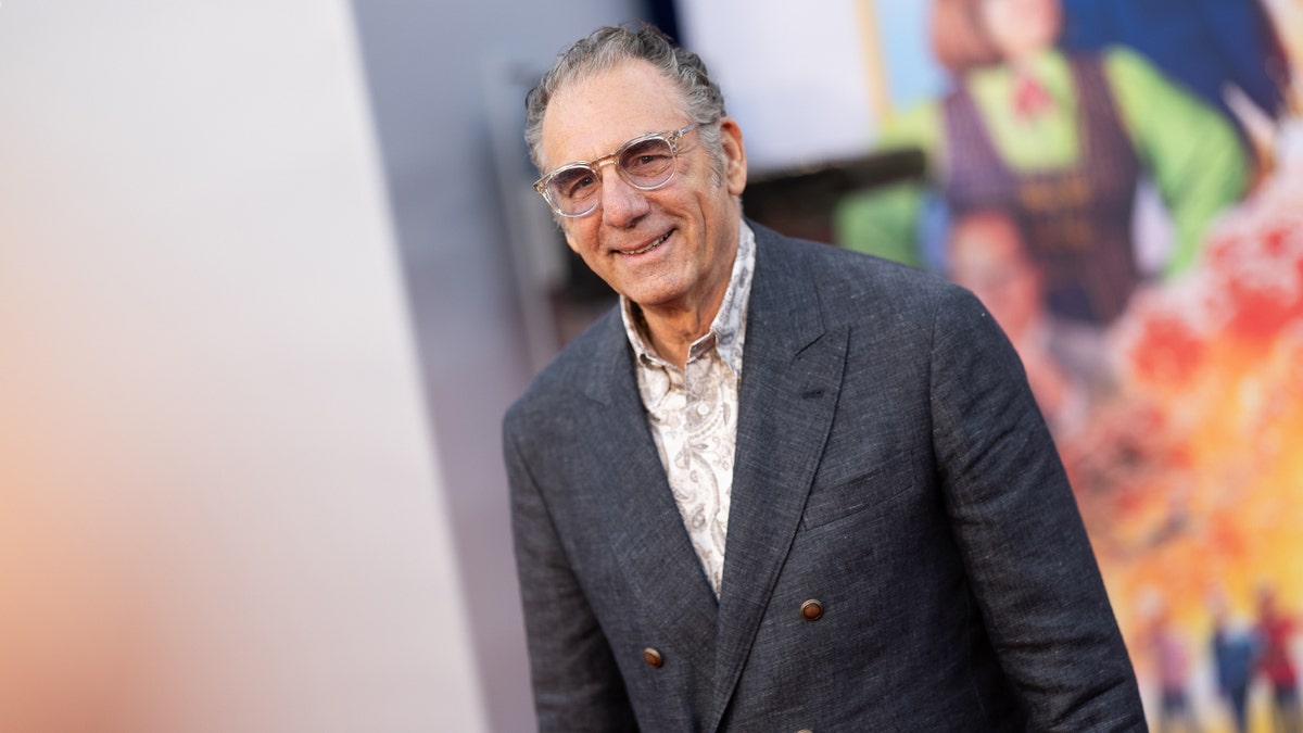 Michael Richards in a dark suit and round glasses smiles on the carpet