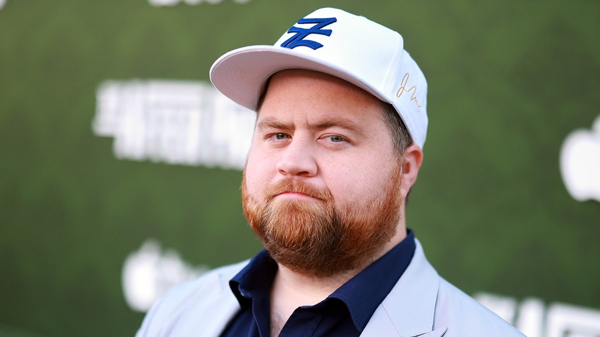 Paul Walter Hauser in a grey suit, navy shirt and white baseball hat stares seriously at the camera