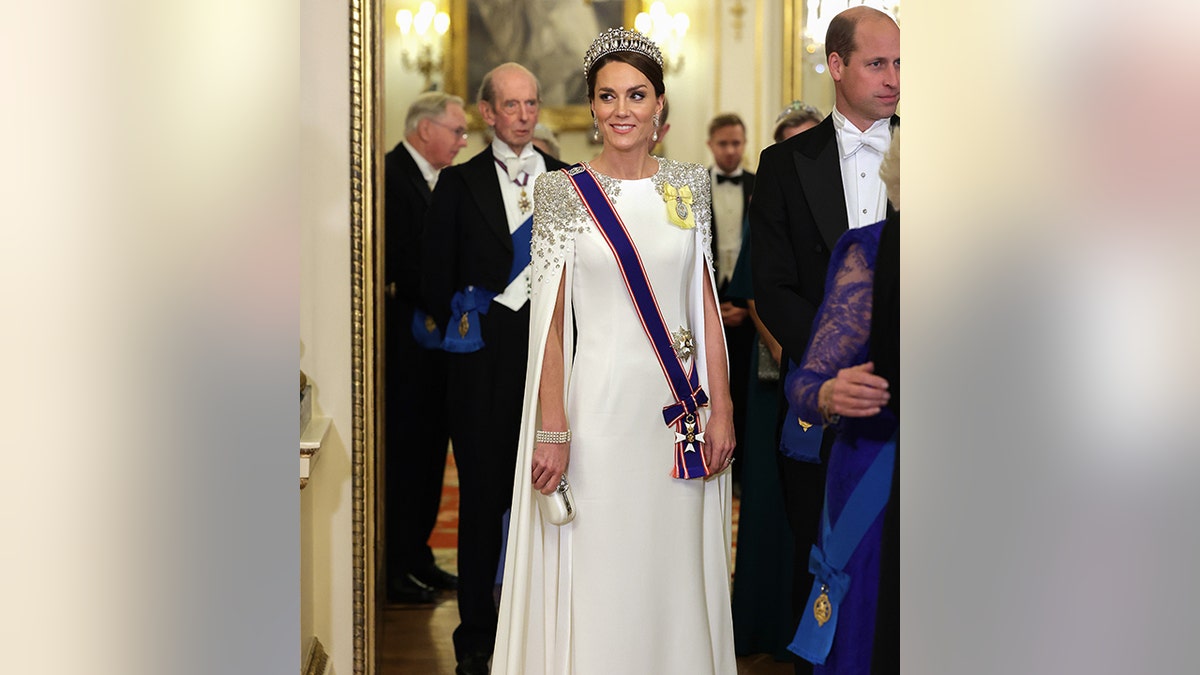 Princess Catherine smiles wearing a long white dress with jewels on the shoulders and a blue sash with Prince William in the background