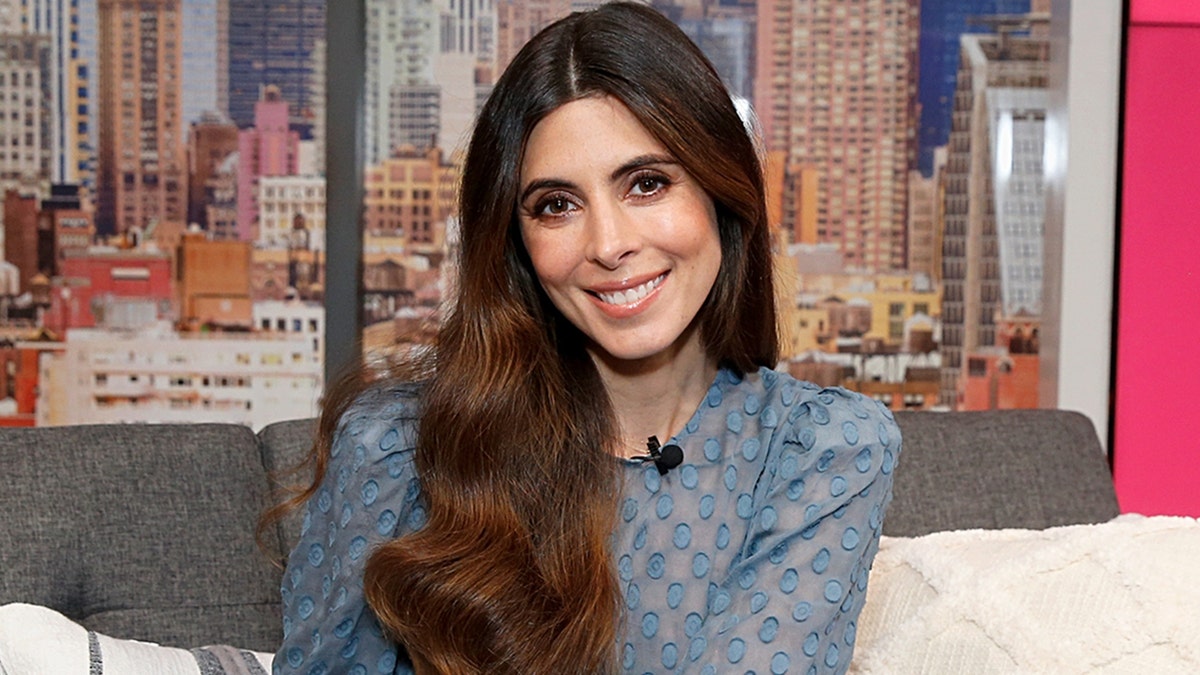 Jamie-Lynn Sigler in a light blue lace dress smiles sitting on a couch
