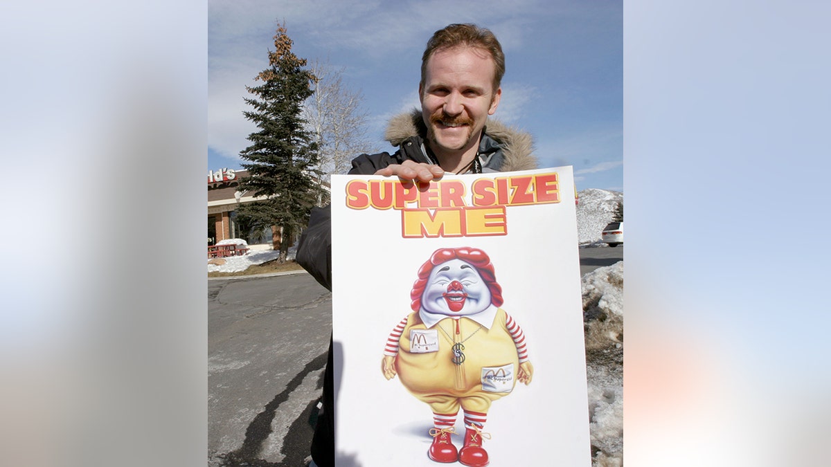 Morgan Spurlock holding a poster that say "Super Size Me" and smiling in a winter coat