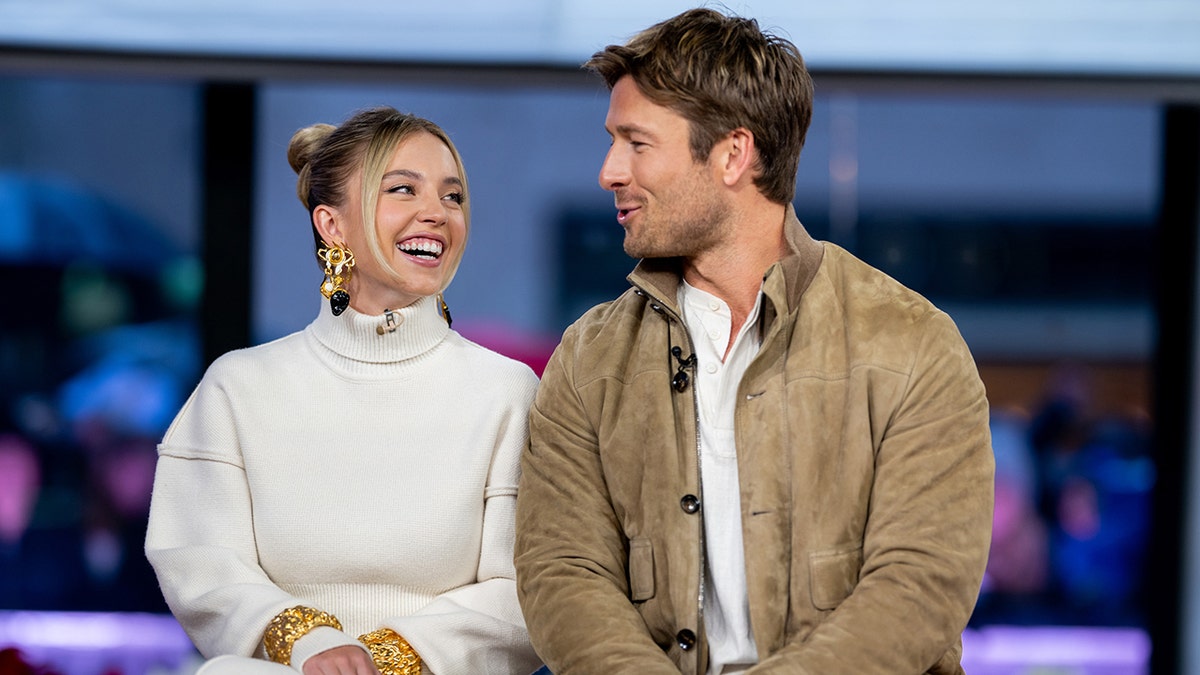 Sydney Sweeney in a cream sweater looks at Glen Powell in tan jacket while on TODAY