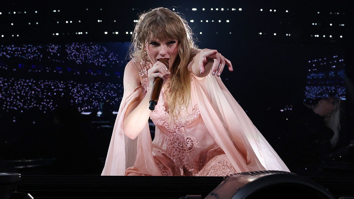 Taylor Swift in a light pink dress points down toward the crowd as she is at the edge of the stage