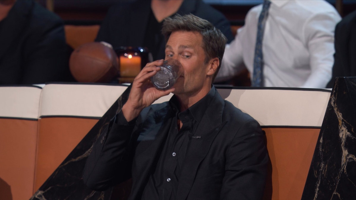 Tom Brady sitting down during his roast taking a sip of his drink