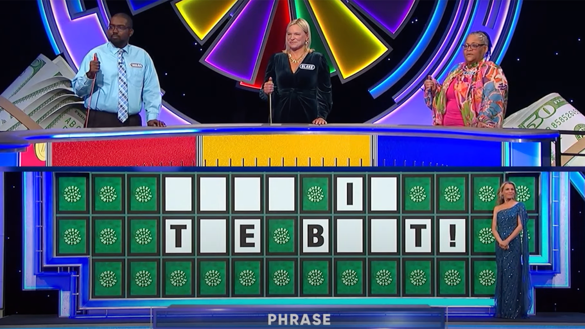 Wide shot of Wheel of Fortune set with three contestants and Vanna White waiting to fill in the blank