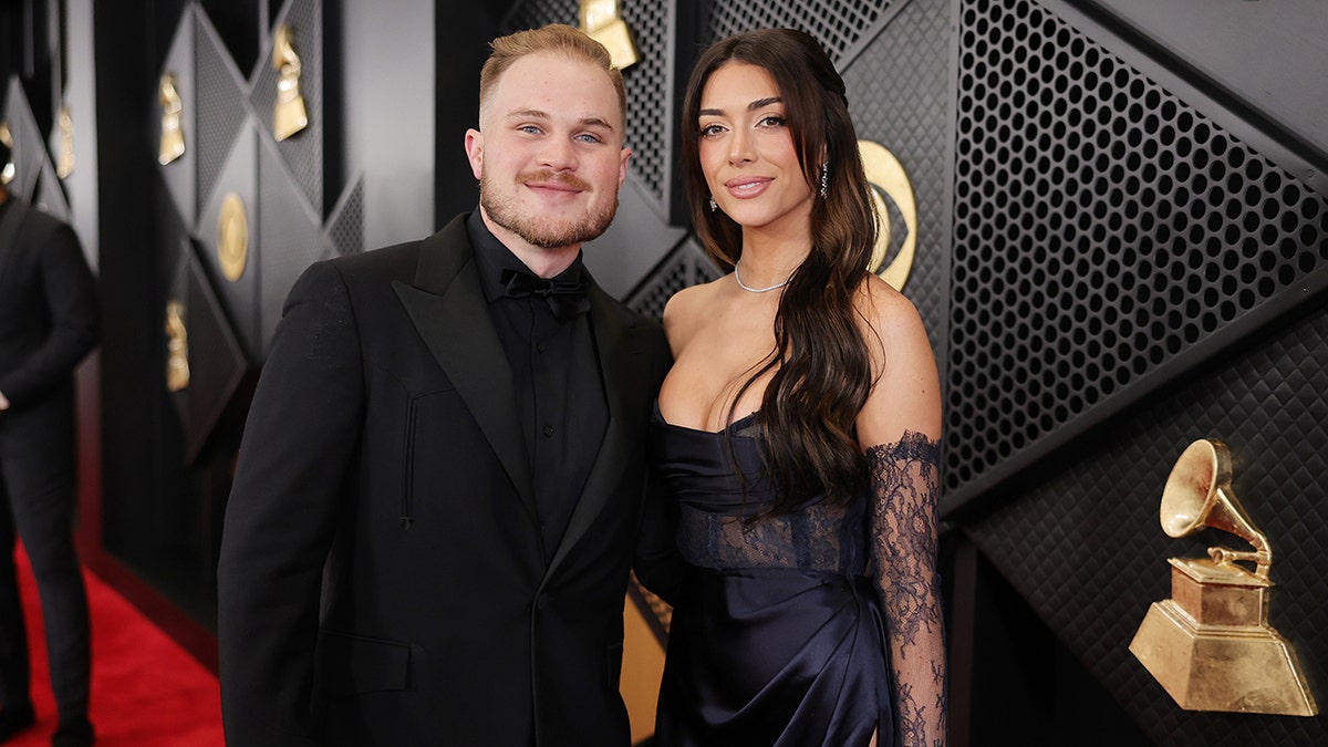Zach Bryan in a black suit stands next to girlfriend Brianna LaPaglia in a blue lace dress at the Grammys