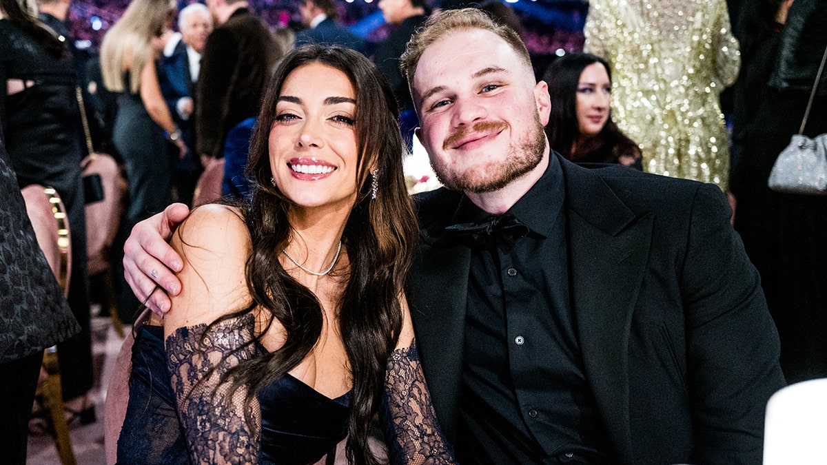 Brianna LaPaglia smiles in a dress with lace sleeves at the Grammys with boyfriend Zach Bryan sitting at a table