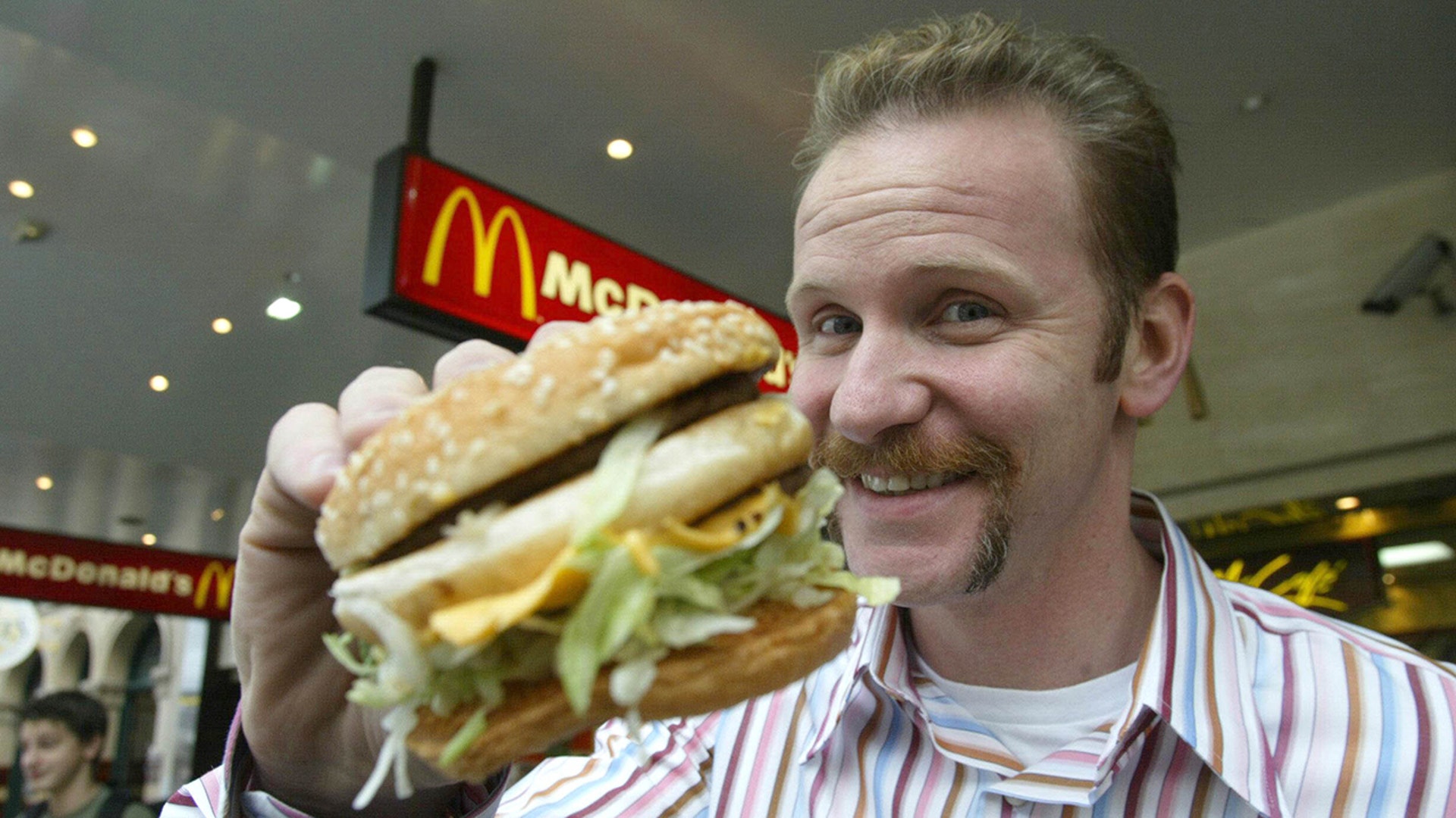 Morgan Spurlock holds up a McDonald's burger to the camera, wearing a striped shirt