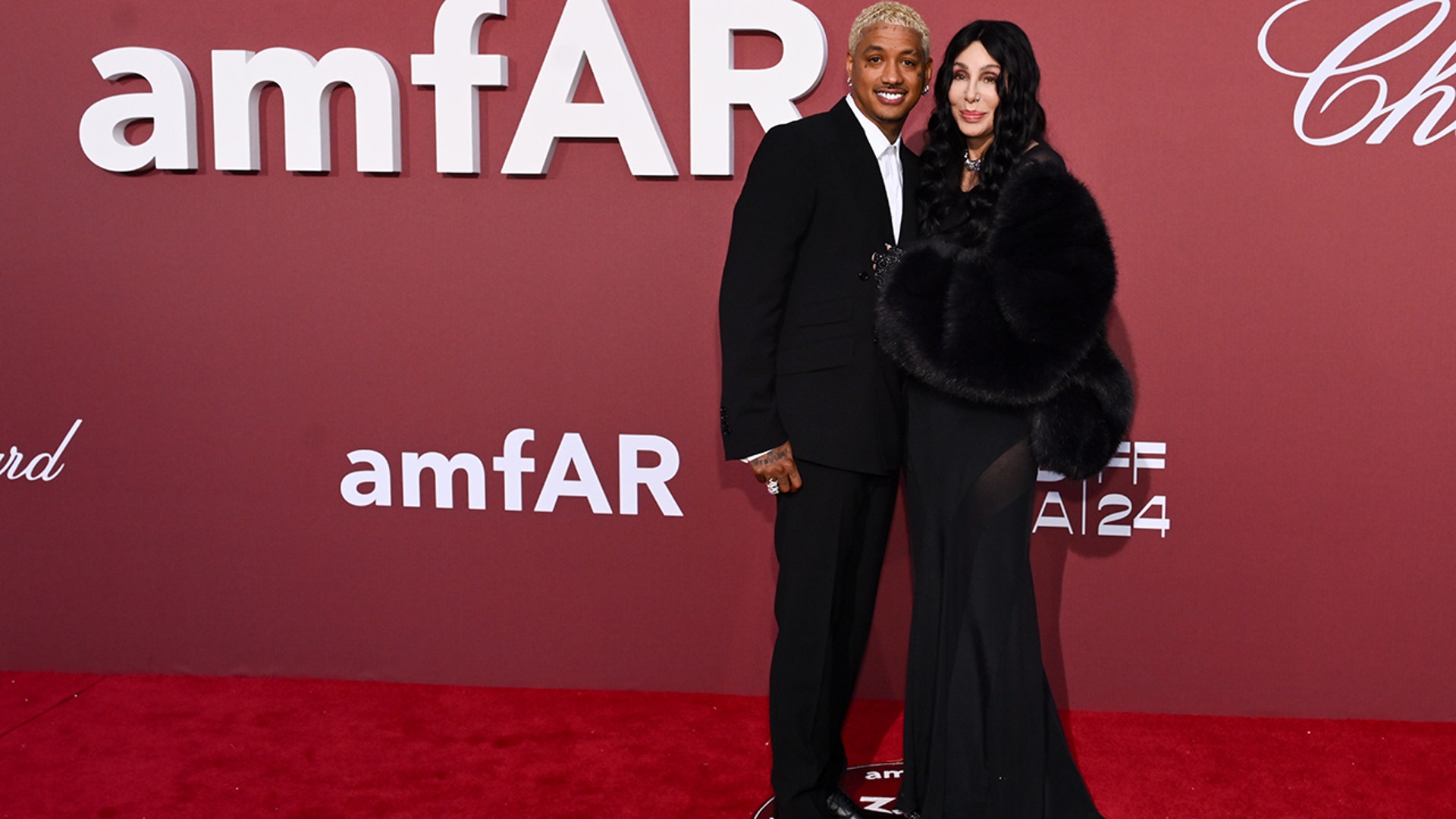 Cher walked the red carpet at the amfAR Gala alongside Alexander Edwards in a black dress with sheer paneling and a black coat, while he wore a black suit.
