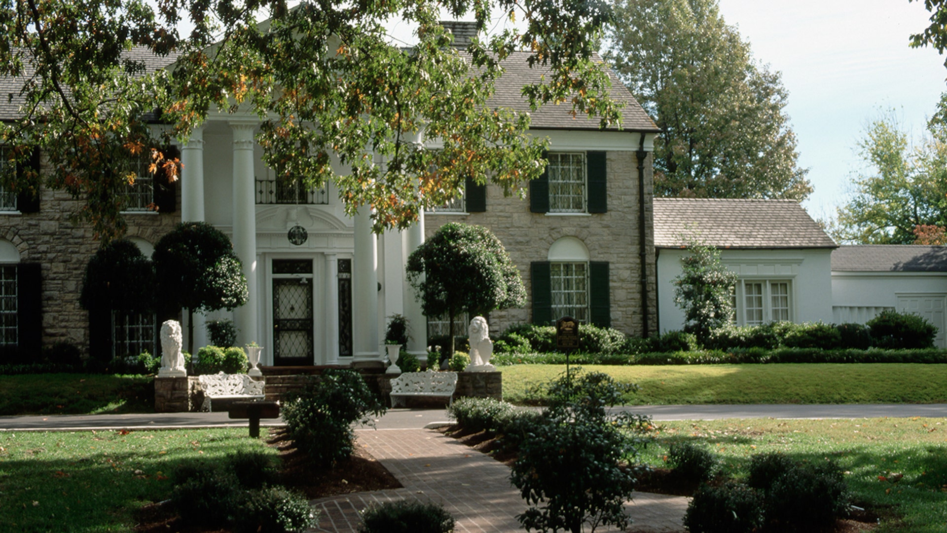 The exterior of Graceland, surrounded by lush green grass and trees, with a brick walkway leading to the front door.