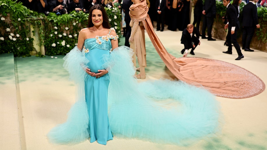 Lea Michele cradles her baby bump wearing a teal gown on the Met Gala red carpet.