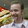Morgan Spurlock holds up a McDonald's burger to the camera, wearing a striped shirt