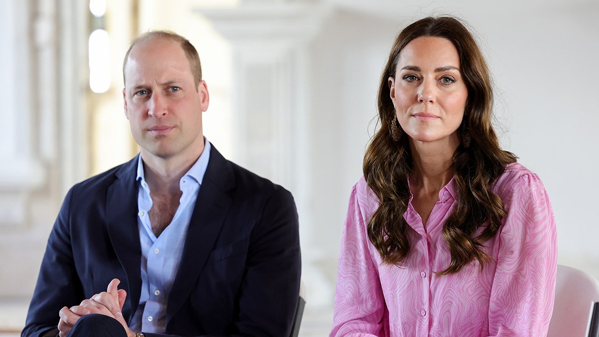 O Príncipe William com um blazer escuro e camisa azul clara parece sério enquanto se senta ao lado de Kate Middleton vestindo uma blusa rosa.