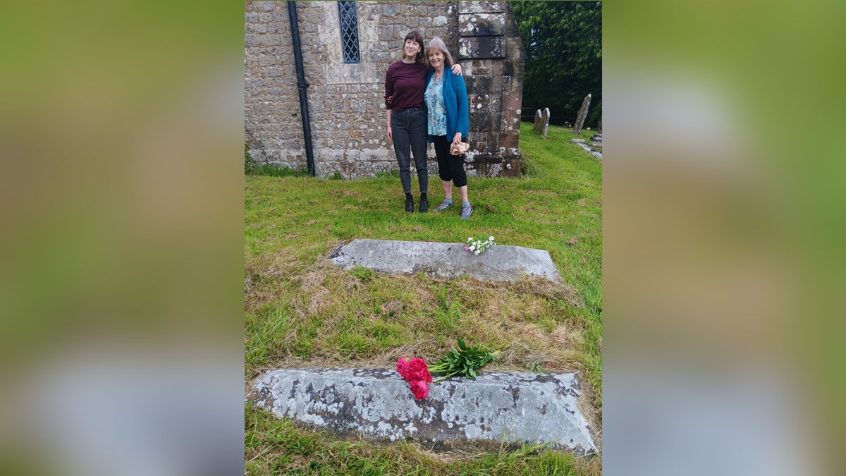 Older woman and younger woman standing at gravesite with flowers.