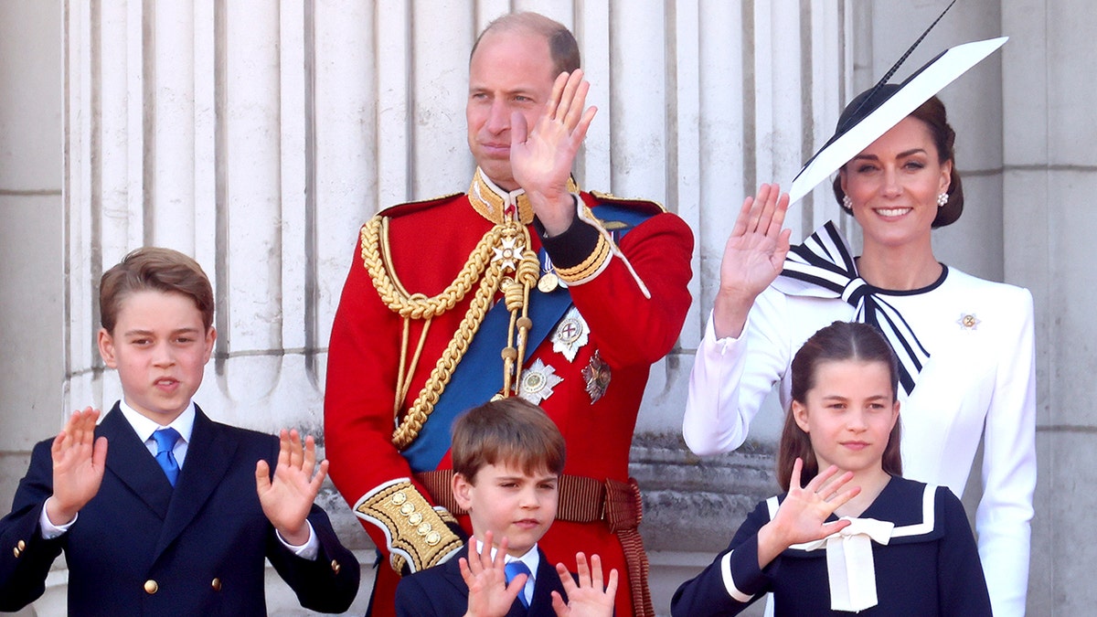 Kate Middleton de branco e o Príncipe William de terno vermelho estão na varanda do Palácio de Buckingham com seus três filhos e acenam para Trooping the Color