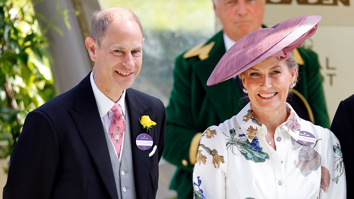 Príncipe Eduardo de terno sorrindo ao lado de sua esposa Sophie em um vestido floral branco com chapéu rosa.