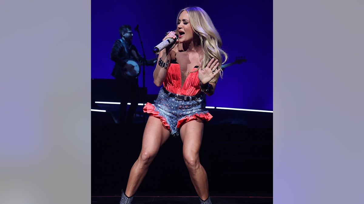 Carrie Underwood con un top bustier rojo y unos shorts vaqueros con ribetes rojos canta apasionadamente al micrófono