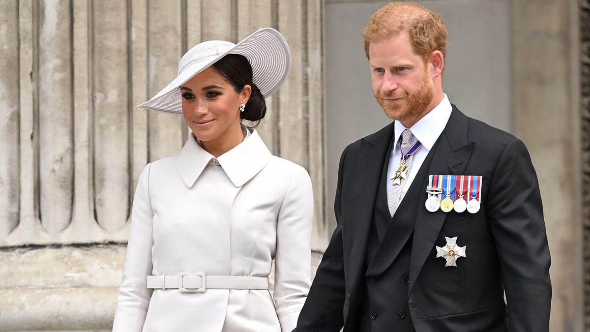 Prince Harry in a dark suit with medals walking alongside Meghan Markle in a white coat dress and matching hat.