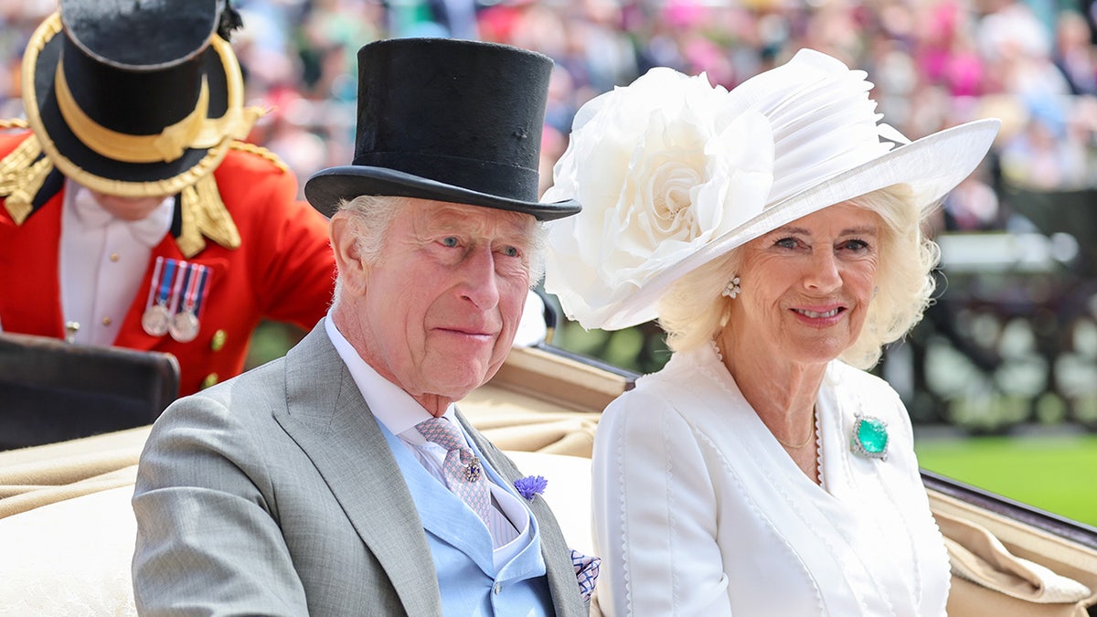 King Charles wearing a grey suit and a black top hat sitting in a carriage next to Queen Camilla wearing all white.