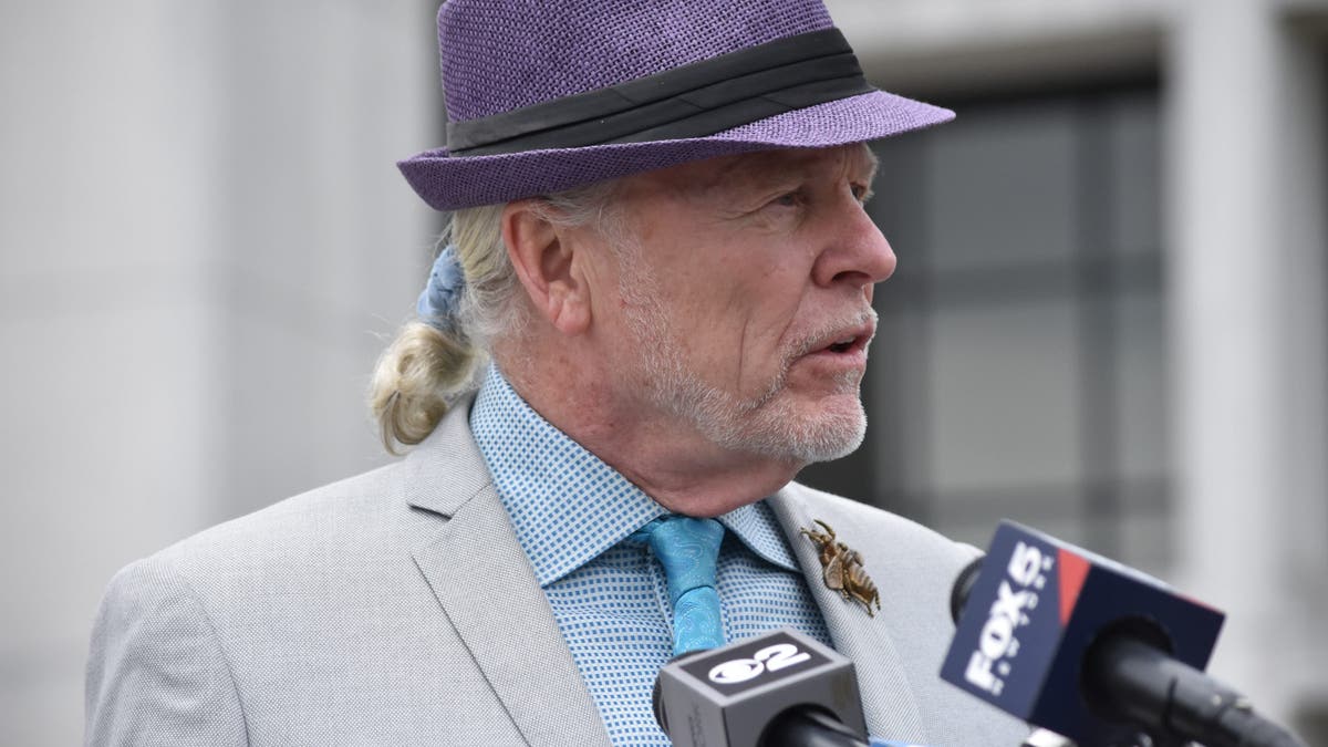 John Ray in a gray suit and purple fedora speaking to reporters